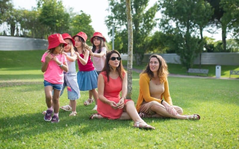 two woman sitting on a grass field while kids are playing