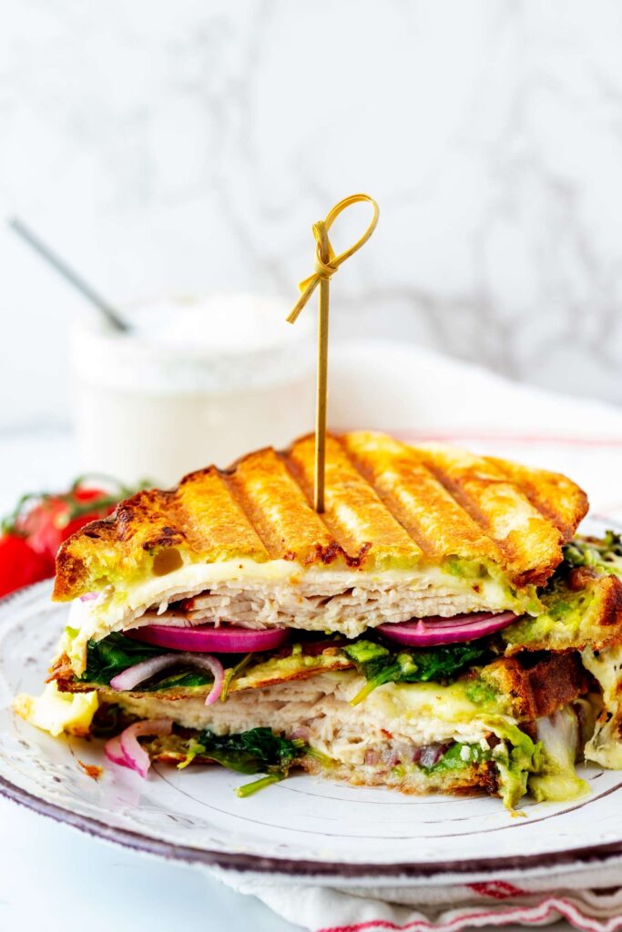 Layered sandwich with turkey, avocado, red onion, and melted cheese filling served on a white plate.